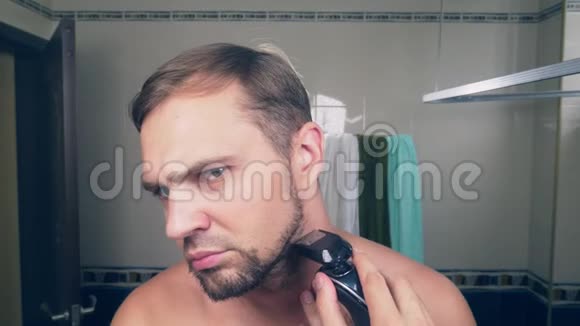 一个英俊的年轻人用电动剃须刀剃胡须站在镜子前的浴室里4k慢视频的预览图