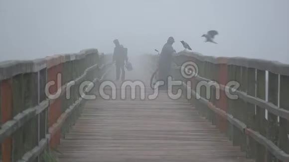 两个渔夫的身影和乌鸦在雾蒙蒙的早晨在古老的木湖大桥上视频的预览图