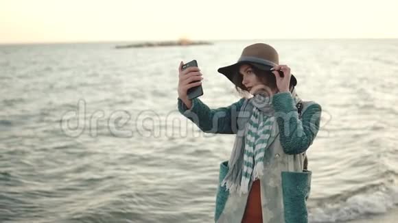 日落时在海滩上自拍的女孩一位身穿秋装的年轻漂亮女子在手机上拍照视频的预览图