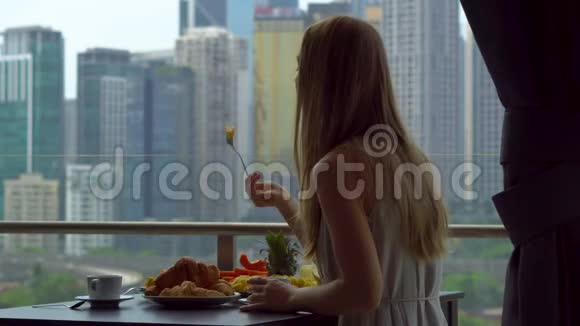 年轻的女家庭教师和一个男孩在阳台上吃早餐俯瞰市中心的摩天大楼视频的预览图