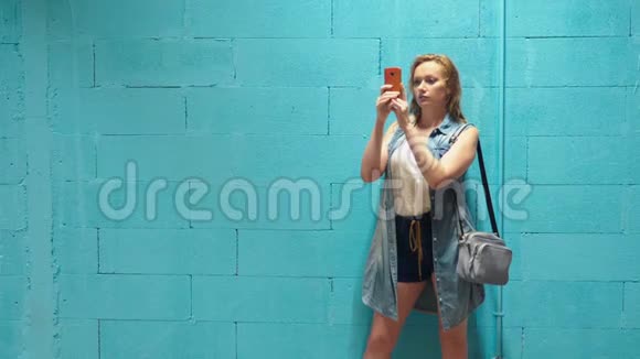 漂亮的金发女孩用红色智能手机对着蓝墙视频的预览图