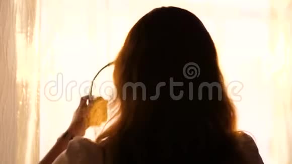 一个年轻的女孩来到窗口戴上耳机听音乐背景模糊与日落女人视频的预览图
