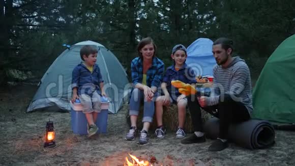 妈妈爸爸和儿子在森林里休息妈妈孩子和爸爸围着火烤玉米全家晚上野餐视频的预览图