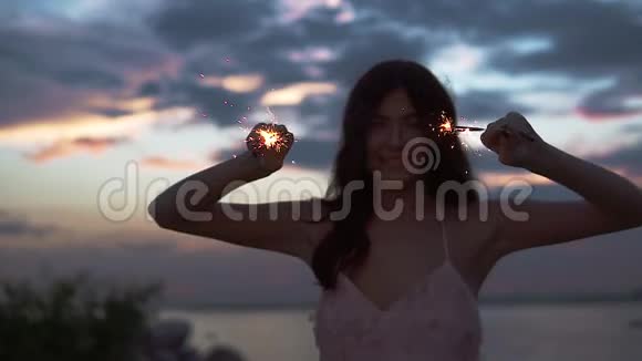 日落时分一个带着火花的迷人女孩的肖像美丽的年轻女子在海滨跳舞慢动作视频的预览图
