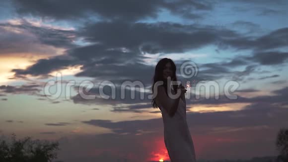 美丽的女孩穿着维切罗裙在日落时在海滩上跳舞一个年轻的女人拿着火花玩得很开心视频的预览图