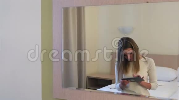 年轻的金发女人在酒店房间的床上坐着平板电脑视频的预览图