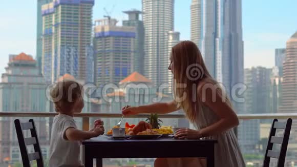 年轻的女家庭教师和一个男孩在阳台上吃早餐俯瞰市中心的摩天大楼视频的预览图
