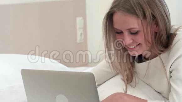 在家里或旅馆房间里躺在床上时戴着笔记本电脑微笑的金发女人视频的预览图