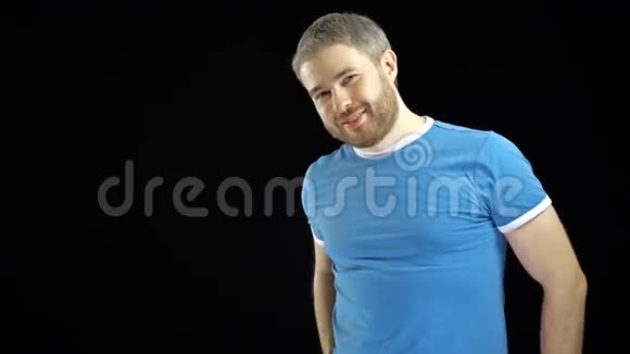 身穿蓝色t恤长胡子的帅哥在黑色背景下对着镜头微笑4K视频视频的预览图