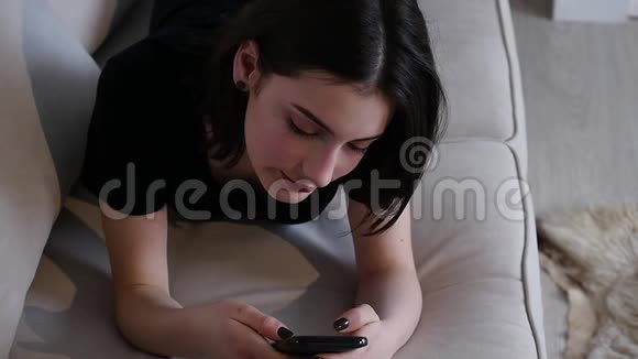 年轻的女人一边用智能手机一边躺在沙发上微笑女孩触摸屏幕浏览互联网视频的预览图