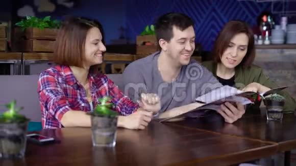 三个微笑的人坐在咖啡馆里选择食物和饮料仔细地看菜单朋友花费视频的预览图