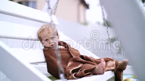 一个一岁的小女孩裹着毛巾脸很脏孤独地坐在花园里的秋千上在花园里视频的预览图
