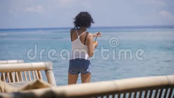 一个留着短发一件T恤和牛仔短裤的女孩站在巴厘岛的海边她喝鸡尾酒视频的预览图