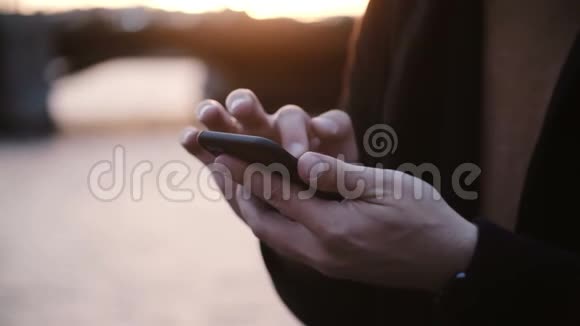 近距离观看男性手拿着智能手机在触摸屏上打字日落时站在河堤上视频的预览图