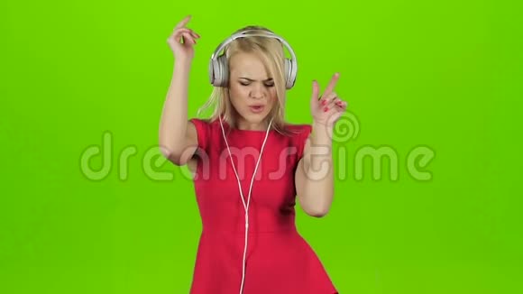 戴着红裙子的金发女孩戴着耳机跳舞戴着绿屏视频的预览图