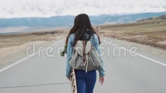 年轻女孩十几岁的嬉皮士用长板在高速公路上行走带着露营背包和运动衫反对视频的预览图