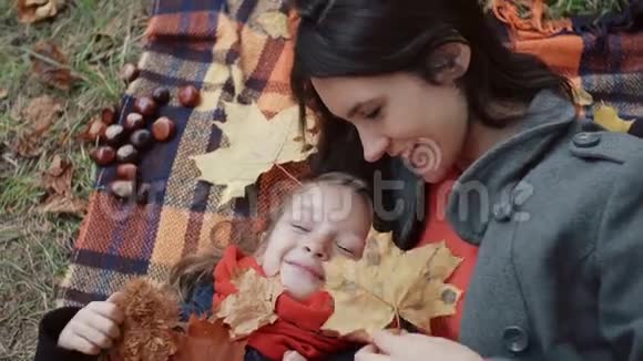 美丽的母亲和她可爱的小女儿躺在格子布上在秋天公园玩枫叶视频的预览图