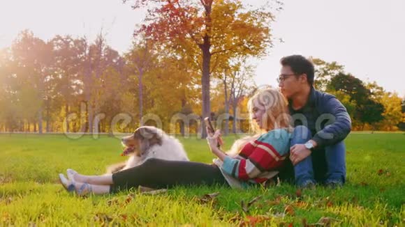 年轻的多民族夫妇和他的狗在公园里放松高加索女人喜欢平板电脑一个韩国男人坐在附近视频的预览图