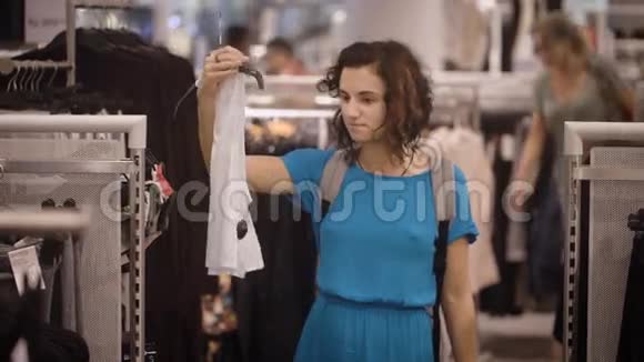 穿着浅蓝色连衣裙的漂亮女孩正在购物并在购买白色T恤或灰色衬衫之间做出选择青年视频的预览图