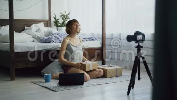 有魅力的年轻女性坐在床边录制关于用dslr摄像机包装礼品盒的视频博客视频的预览图