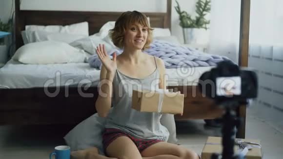 一位开朗的女士坐在床边录制关于用dslr相机包装礼品盒的视频博客视频的预览图