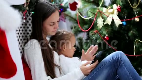 年轻的母女在圣诞装饰的室内使用智能手机视频的预览图