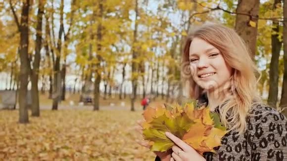 一个年轻的女孩高兴地在相机上摆着一束五颜六色的叶子秋天在城市公园散步视频的预览图
