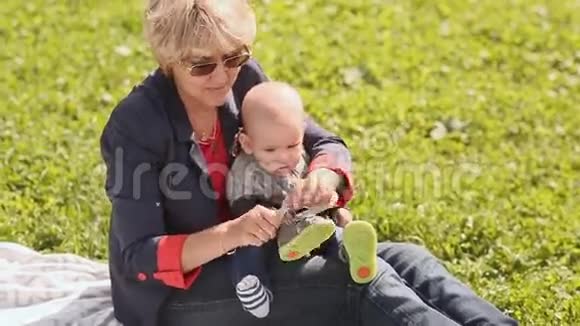 年轻的祖母坐在绿色的草鞋凉鞋上宝宝的脚在夏天视频的预览图