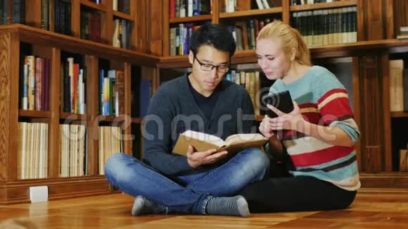 朋友们坐在图书馆的地板上一个男人拿着一本打开的书一个女人用了一块平板电脑视频的预览图