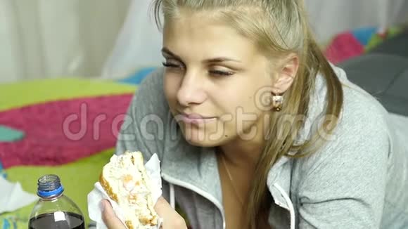 很高兴的女人在她的房间里的床上吃快餐汉堡和汽水很垃圾的食物慢动作视频的预览图