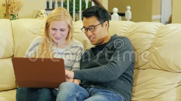 跨国夫妇在家放松坐在沙发上使用笔记本电脑视频的预览图