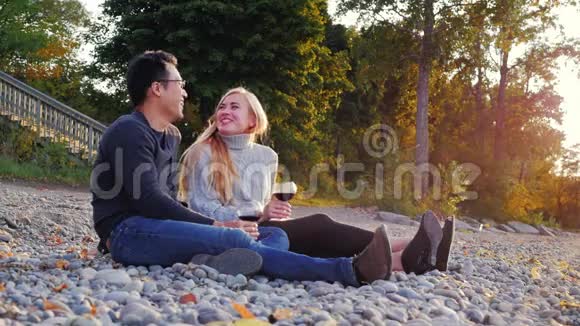 年轻夫妇在鹅卵石海滩上休息他们喝杯酒亚洲男人和高加索女人视频的预览图