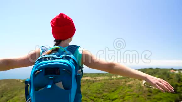 背着一个背包的女人正把手臂向两边旋转站在海洋上的一座小山上视频的预览图
