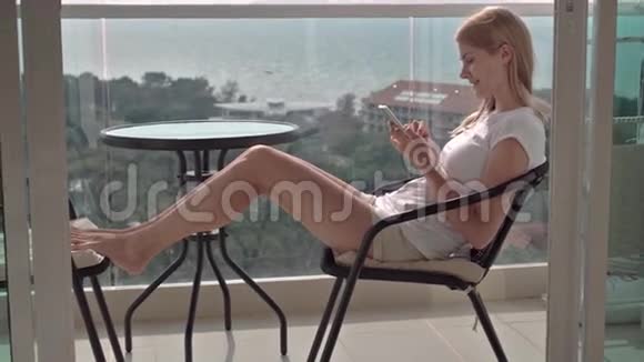坐在阳台椅子上浏览互联网的美丽迷人的女人与她的朋友交流视频的预览图