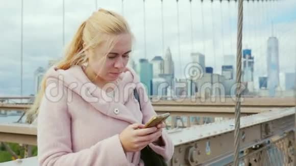 年轻女子在你的智能手机上打短信站在纽约布鲁克林大桥的栏杆旁视频的预览图