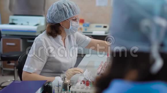 研究助理穿着白色实验室外套戴着防护眼镜戴着面具戴着帽子和手套端坐在桌子旁视频的预览图