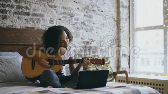 好奇的非裔美国少女集中精力在家里坐在床上学习用笔记本电脑弹吉他视频的预览图
