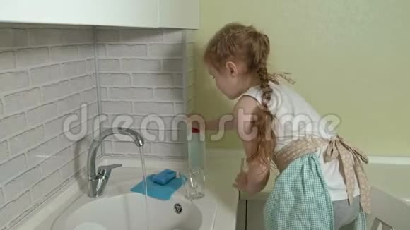 漂亮的小女孩穿着围裙在明亮的厨房里洗了一个杯子帮助父母视频的预览图