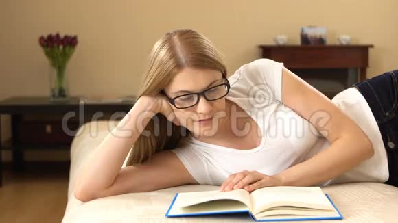 年轻漂亮的女人穿着白色的t恤和眼镜轻松地躺在沙发上享受着看书的乐趣视频的预览图