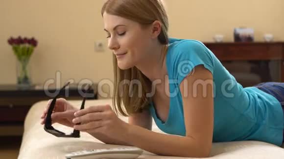 美丽迷人的年轻女子穿着蓝色t恤躺在沙发上穿着3D眼镜看电视视频的预览图