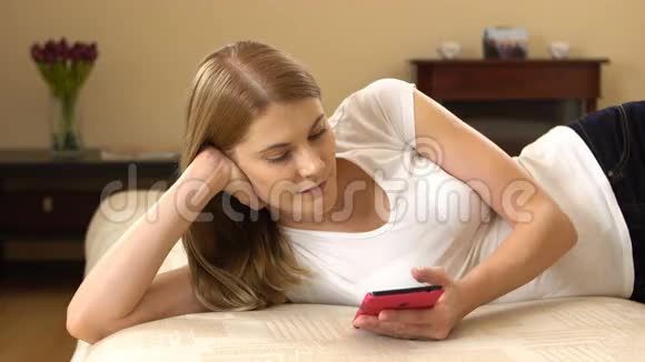 漂亮的年轻女人穿着白色的t恤粉红色的智能手机躺在沙发上浏览互联网微笑视频的预览图