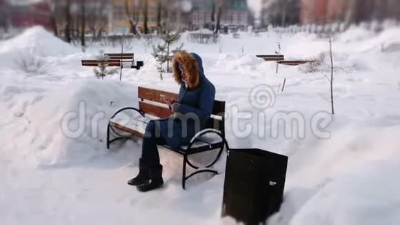 白天在下雪的天气里女人坐在长椅上在冬天的城市公园里浏览手机视频的预览图