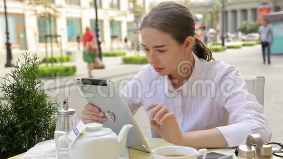聪明的女学生喝咖啡看着她的平板电脑在街上的自助餐厅打电话给她的手机视频的预览图