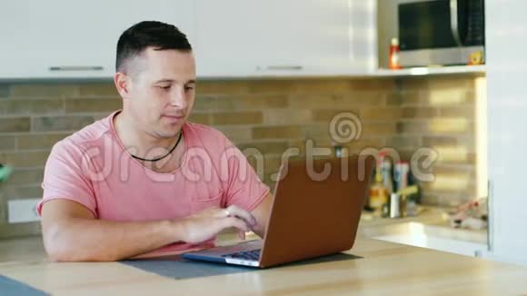 年轻有魅力的男人在家里的厨房里用笔记本电脑工作视频的预览图