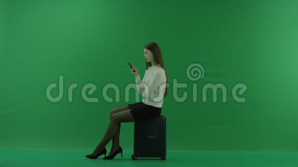 女孩看着左边坐在手提箱上看着绿色屏幕上的电话视频的预览图