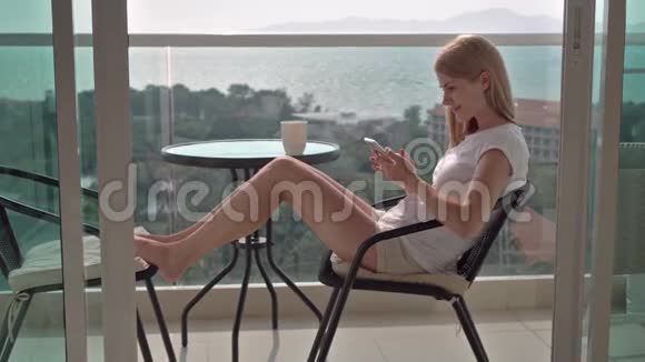 坐在阳台椅子上浏览互联网的美丽迷人的女人与她的朋友交流视频的预览图