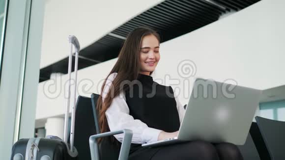 机场年轻女乘客在候机大厅坐着智能手机和笔记本电脑等待她的航班工作视频的预览图