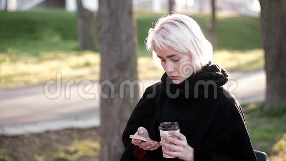 外面的金发女孩在智能手机和喝咖啡的手里智能手机上的文字类型视频的预览图