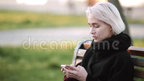 金发女孩在外面看着长凳上的智能手机在智能手机上玩一个活跃的3D游戏微笑视频的预览图