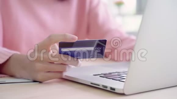 美丽的亚洲女人用电脑或笔记本电脑通过信用卡购买网上购物而穿毛衣坐在桌子上视频的预览图